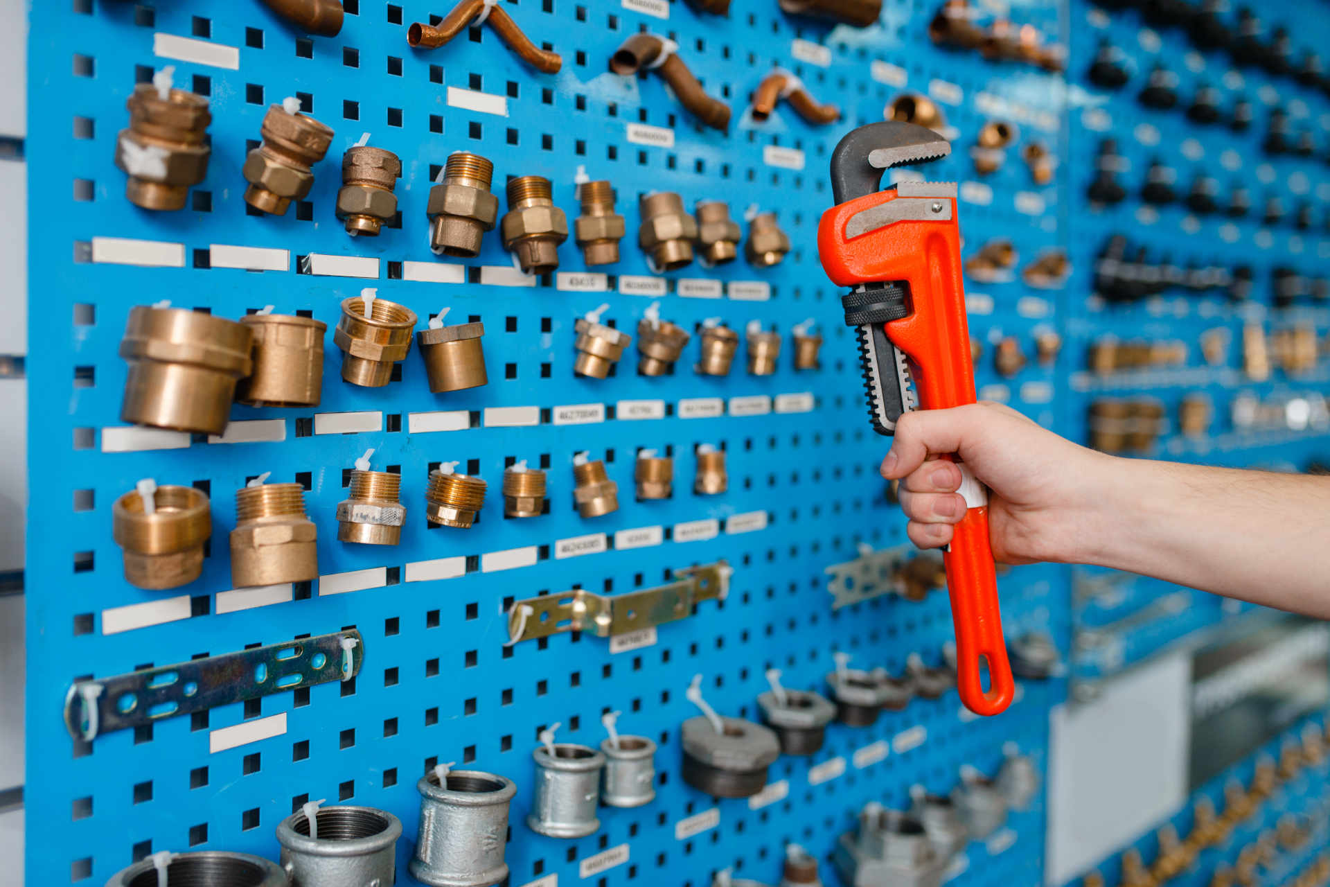 Mano sujetando una llave de tubo roja frente a una pared azul con múltiples accesorios de plomería organizados.